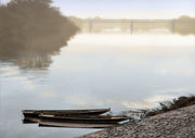 Two wooden rowboats moored at a cobblestone riverbank, fog obscuring a distant bridge and treeline