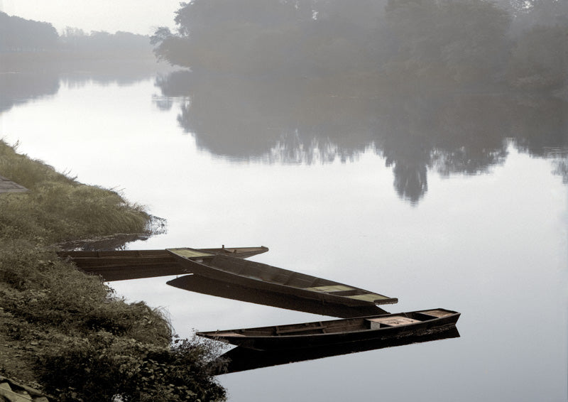 Three wooden boats moored on a still, foggy river with a misty tree-lined bank