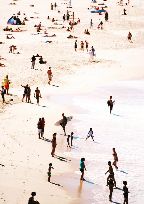 Aerial view of a sunlit beach with scattered people, long shadows and pale turquoise shoreline.