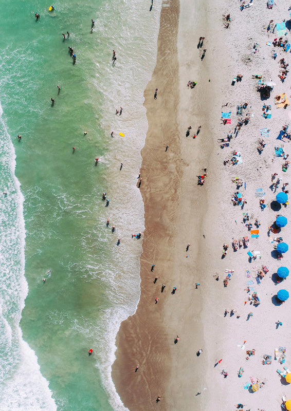 Aerial photo of teal ocean waves meeting tan sand with swimmers and blue beach umbrellas
