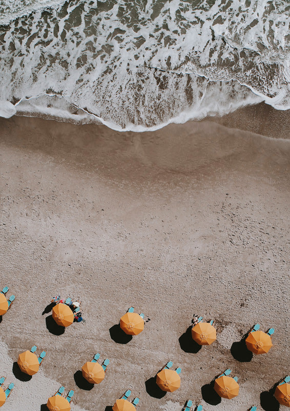 Aerial photo of orange beach umbrellas and turquoise loungers on sand beside foamy ocean waves