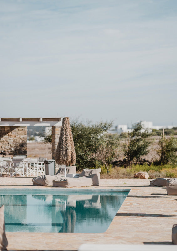 Rectangular pool beside a stone pergola with white chairs and olive trees under a pale sky