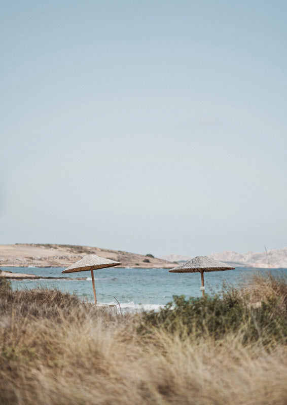 Two straw beach umbrellas on a grassy dune beside calm blue water under a pale sky