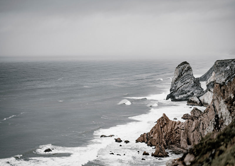 Grey moody photograph of rugged coastal cliffs with waves crashing below foggy sky