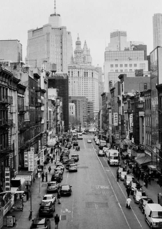 Black and white photo of a busy New York street with storefronts, fire escapes and skyscrapers beyond