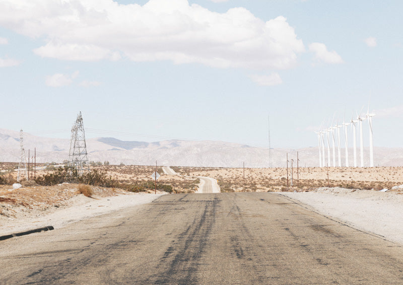 Desert road leading to distant hills with a power pylon and wind turbines under a pale sky