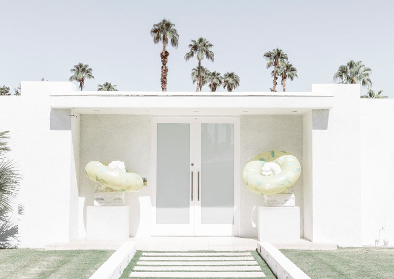 White mid-century building with palm trees, symmetrical doors and pale yellow floats on plinths