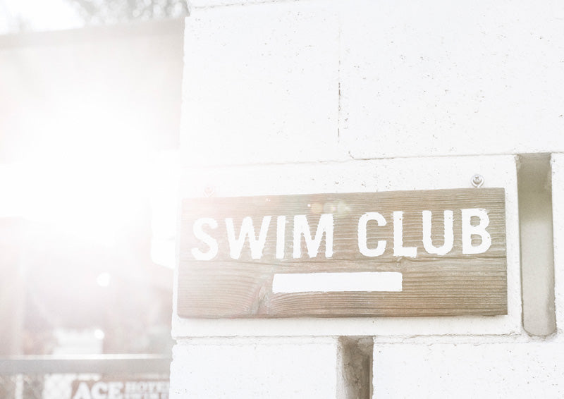 Weathered wood sign reading Swim Club on a white brick wall with sun flare