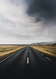 Straight road leading to a horizon under storm clouds, flanked by golden fields and mountains.
