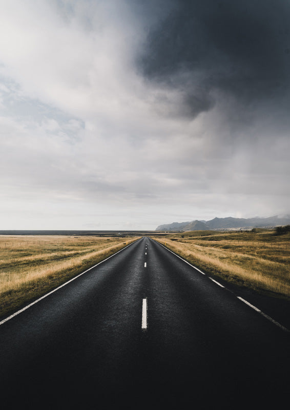 Straight road leading to a horizon under storm clouds, flanked by golden fields and mountains.