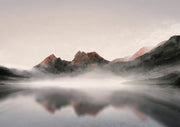 Misty mountain range with rust-toned peaks reflected in calm water beneath a pale sky