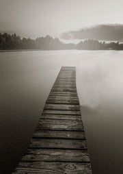 Sepia-toned photograph of a wooden jetty extending into a calm lake, framed by trees and clouds.