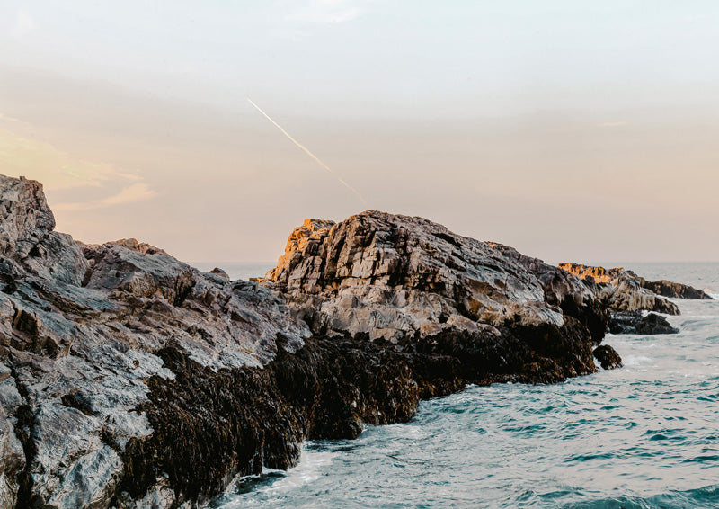 Rocky coastline with jagged cliffs, teal ocean waves, and a pastel sunset sky with a faint jet trail
