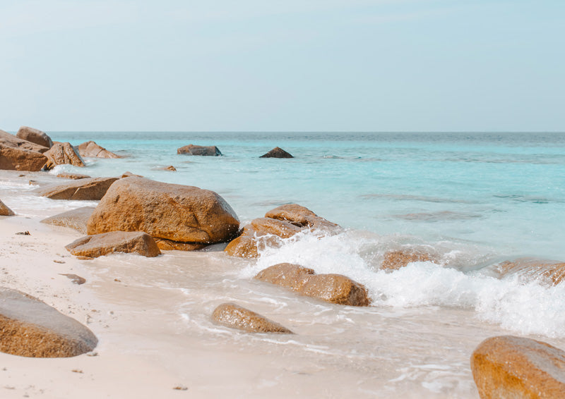 Turquoise ocean water and terracotta boulders on a sandy beach with white surf and pale sky
