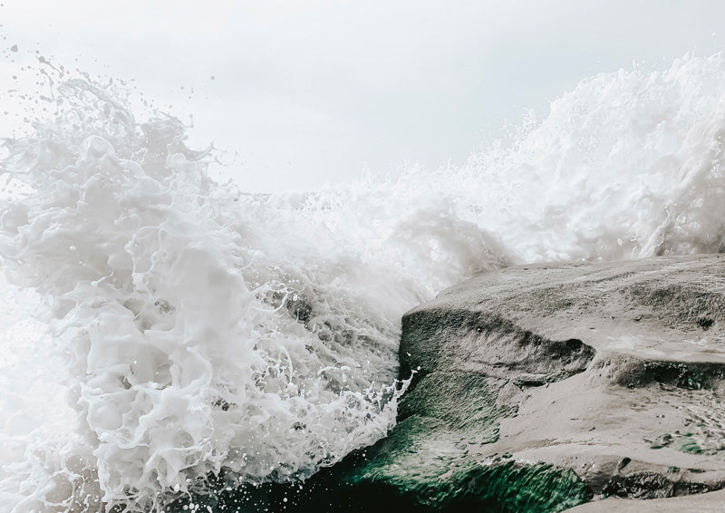 Photograph of a wave crashing over dark rock, white spray against a pale sky and green water below.