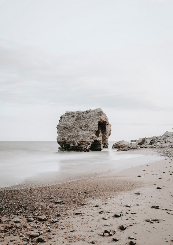 Weathered rock formation on a beach shoreline with soft, misty tide and grey sky