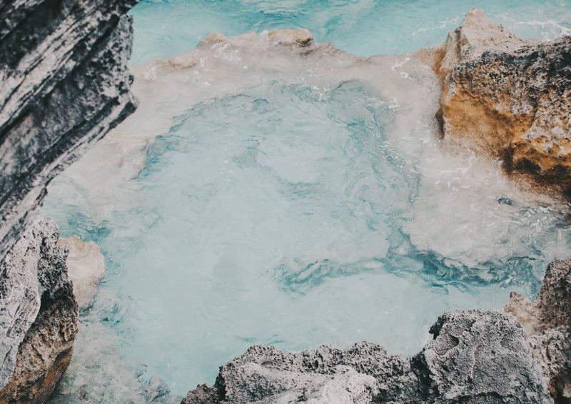 Aerial view of a turquoise rock pool surrounded by grey and rust-coloured rocks with white foam