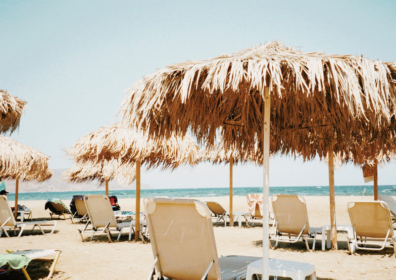 Thatched beach umbrellas and sun loungers on sand with sea and pale sky behind