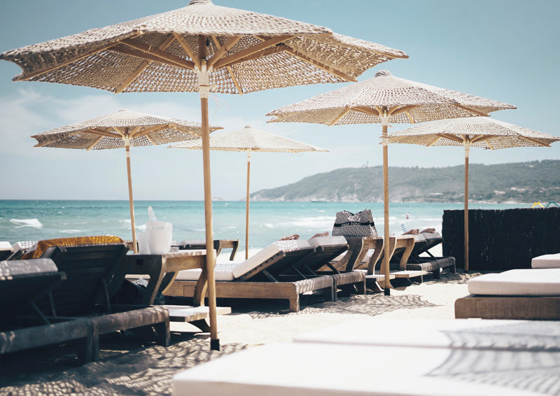 Woven beach umbrellas and timber loungers on sand facing a turquoise sea and coastal headland