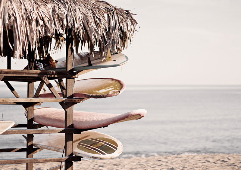 Weathered surfboards stacked beneath a thatched roof beside the ocean and sand.