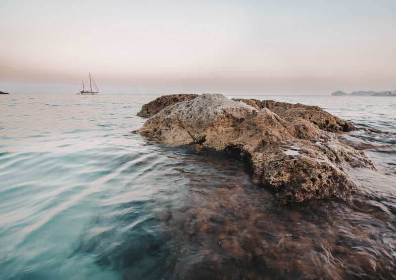 Rock formation in calm turquoise ocean water with a distant sailboat and soft peach sunset sky
