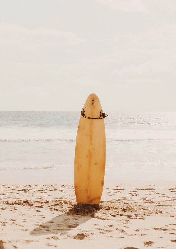 Yellow surfboard standing upright in sand facing the ocean under a pale sky