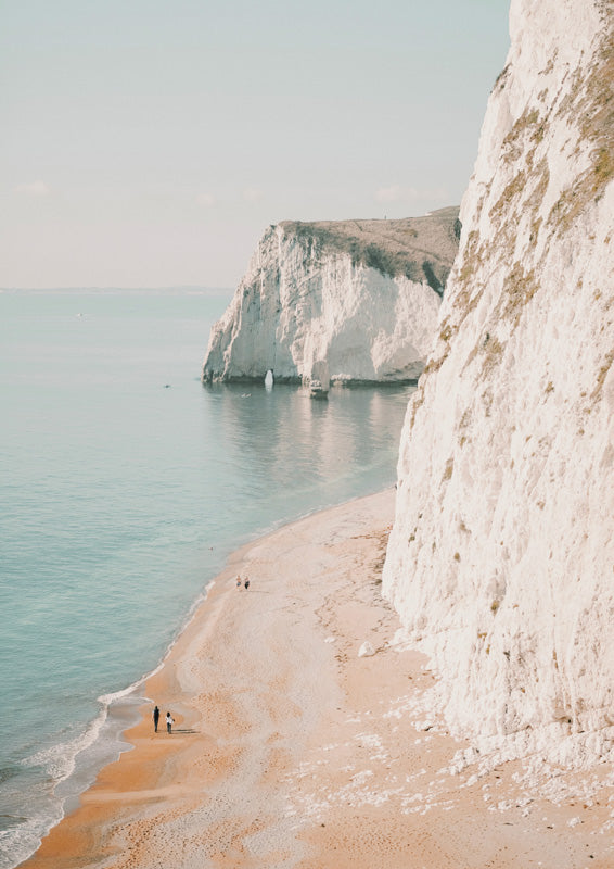 Chalky white cliffs above a turquoise sea, with two figures walking along a sandy beach.
