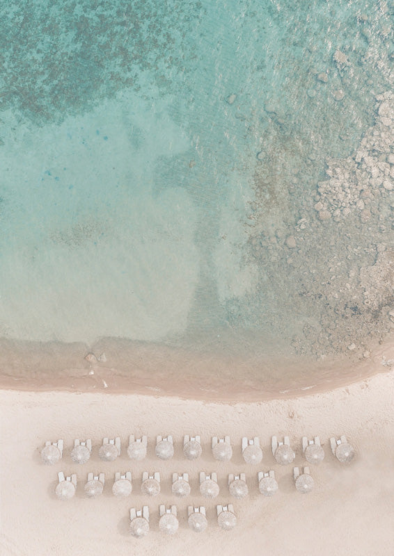 Aerial view of teal ocean water, rocky shallows and rows of white beach umbrellas on sand