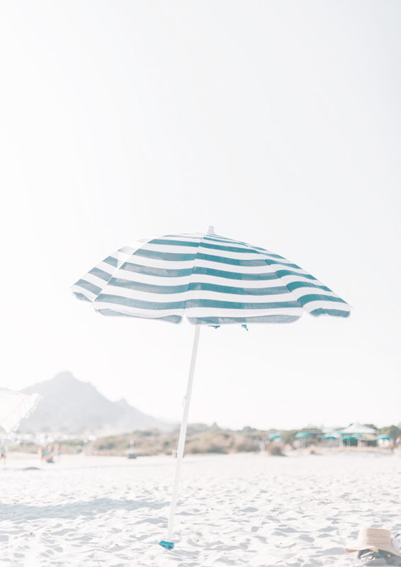 Teal and white striped beach umbrella on sand with hazy hills and buildings in the background