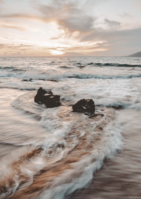 Long-exposure photo of ocean waves swirling around dark rocks under a peach and grey sunset sky