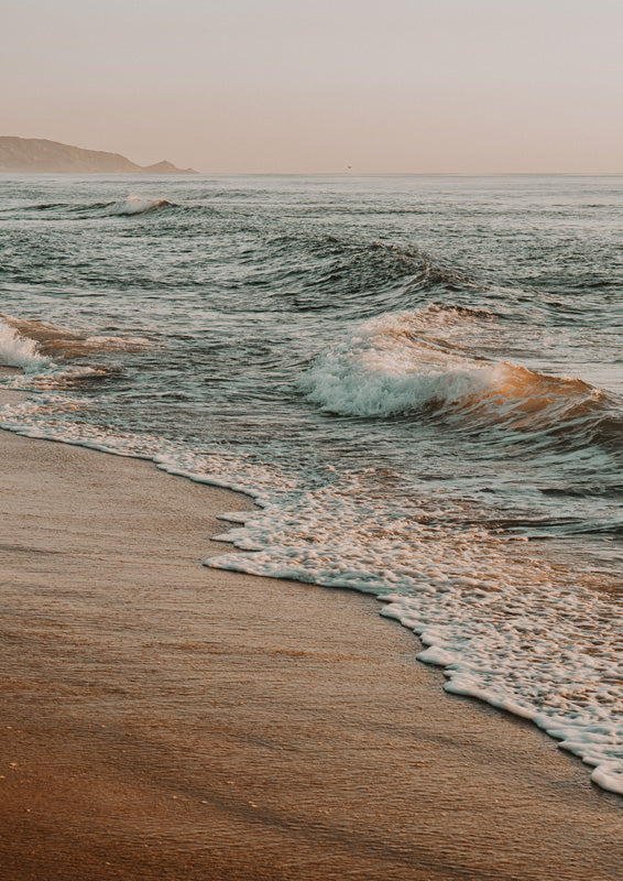 Ocean waves breaking on a sandy shore at sunset with peach and teal tones