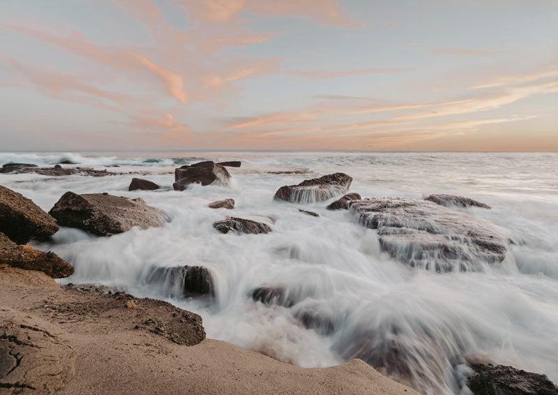 Long exposure photo of ocean waves washing over rocks at sunset with peach sky