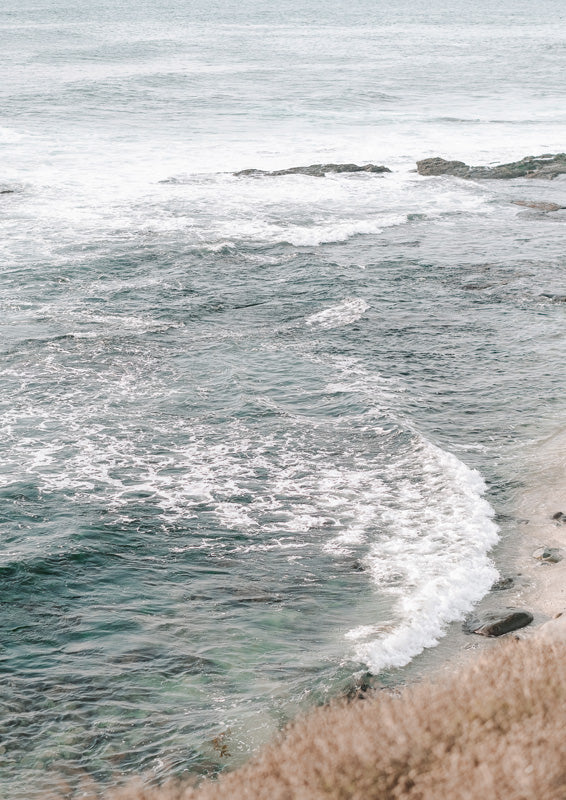 Aerial photograph of ocean waves breaking over rocks near a sandy shoreline