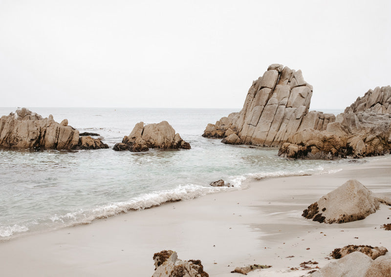 Rocky granite boulders on a sandy beach with calm shallow water under an overcast sky