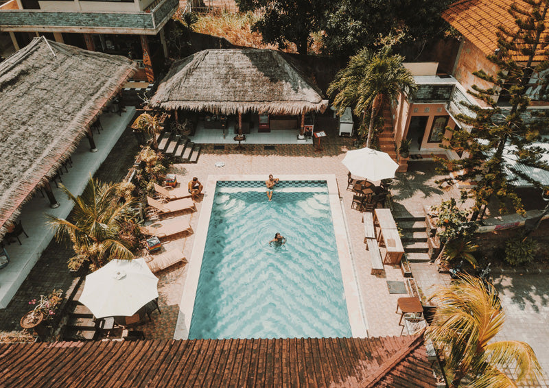 Aerial view of a resort pool with swimmers, palm trees and thatched roof buildings