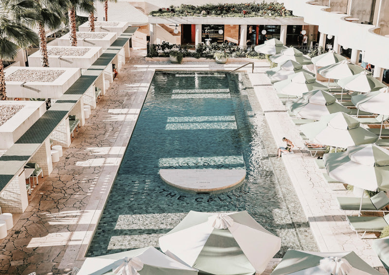 Aerial view of a resort pool with teal mosaic tiling, cream umbrellas and palm trees.