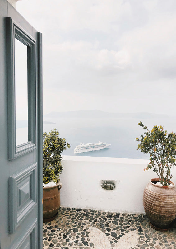 Open blue door framing a sea view with a distant ship, terracotta pots and pebble floor
