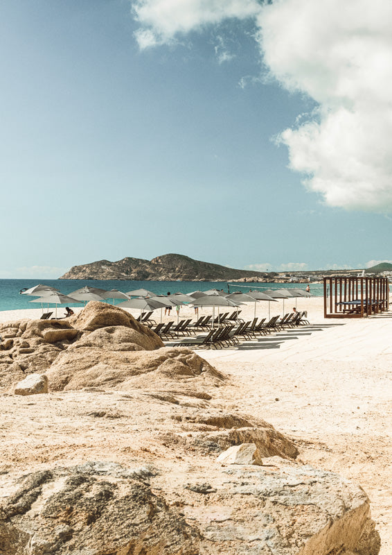 Beach with rocks, umbrellas, loungers, turquoise sea and distant headland under blue sky.