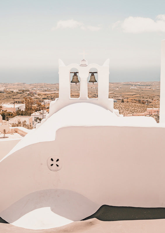 White Santorini bell tower with twin bells and cross against pale sky and arid landscape