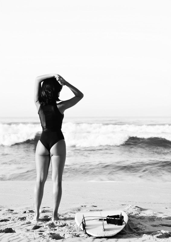 Monochrome photo of a woman facing the ocean beside a surfboard on the beach