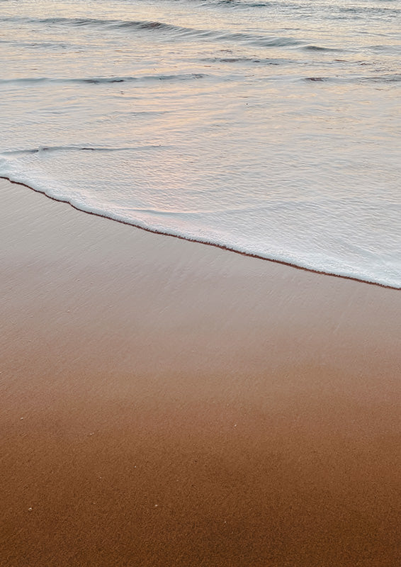 Close-up of a wave meeting a sandy beach at sunset, with soft peach and tan tones.