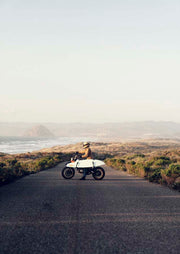 Rider on a white motorcycle with a surfboard on an empty coastal road overlooking the ocean