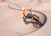 Aerial view of a BMX rider in a concrete skate park bowl, casting a shadow on the pale surface.