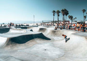 Skater in a concrete skate park bowl with palm trees and beach crowd in the background