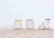 Three striped deck chairs in tan, blue and grey stand on a pebble beach facing the sea