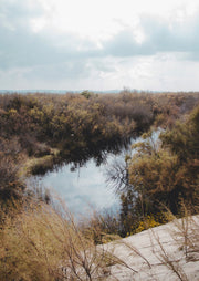Still creek reflecting cloudy sky, surrounded by scrubland and sandy banks