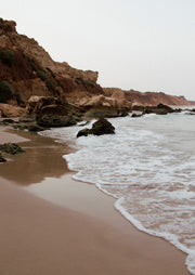 Sandstone cliffs along a beach with gentle waves washing onto wet sand under a pale sky.