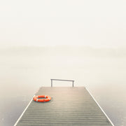 Orange lifebuoy on a wooden pier extending into fog-covered water with a faint treeline