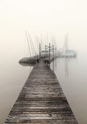 Weathered wooden pier leading to moored sailboats in thick harbor fog