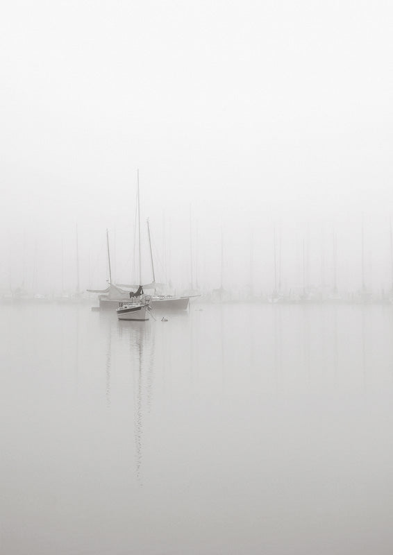 Black and white photo of sailboats moored in a foggy harbour with still reflective water
